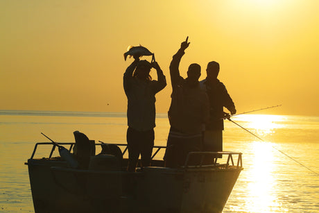 Three people on a boat at sunset, with one person holding a fish.